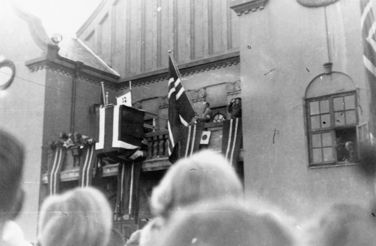 Feiring av freden 1945. Festiviteten pyntet med flagg. Tilskuere følger med. Trolig fra hyllesten av "Tysklandsfangene" som ankom Haugesund 28. mai. Den dagen ble balkongen på festiviteten pyntet akkurat på denne måten. Her; mens de venter på hedersgjestene.