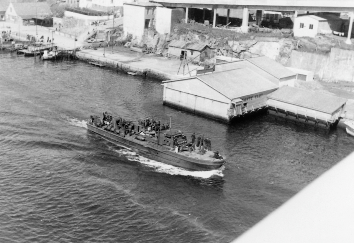 Feiring av freden 1945. Marinefartøy med mannskap på tur sørover sundet. Kaien og søyler til Risøybroen i bakgrunnen. Foto tatt fra Risøybroen. Kan være fra 7. juni 1945 (kongedagen) da Haugesud fikk et stort flåtebeøk.