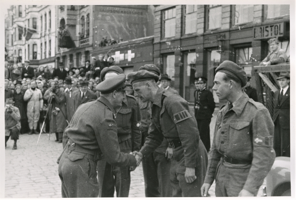 Milorgs distriksjef Alf Skare ønsker velkommen på indre kai den 14. mai 1945. På høyre side står områdesjef Johannes Skogland. I bakgrunnen ser en hotel Brisol. Bildetaking og mulig filming til venstre i bildet. I bakgrunnen ser vi politikonstabler og en deligasjon fra England (menn med hatt og frakk). Mange fremmøtte haugesundere og deler av Hjemmestyrkene ser vi også til venstre i bildet.