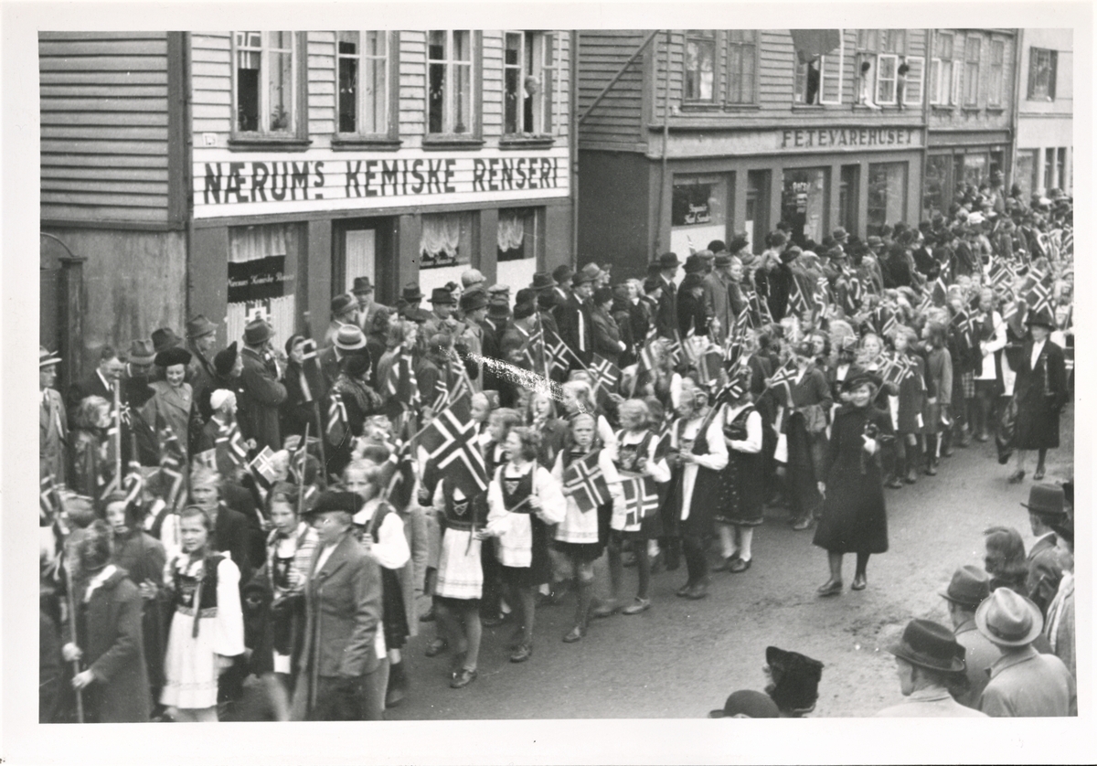 Barnetoget 17. mai 1945 marsjerer gjennom Strandgata like nord for Risøybroen. Tilskuere langs gaten på begge sider og bakerst i bildet ser vi Nærums Kemiske Renseri og Fetevarehuset.