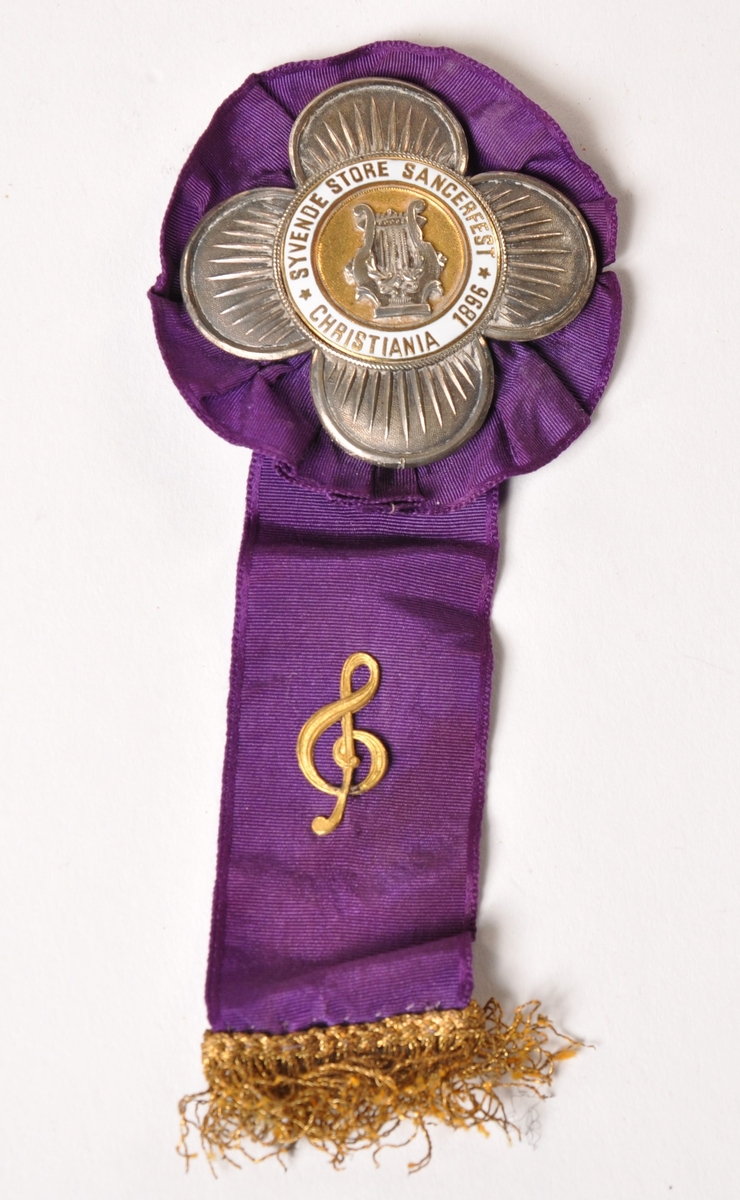 Blomsterformet medalje i sølv med hvit emalje montert på lilla bånd med rosett.  Og g-nøkkel på båndet. Motiv: Harpe på gullbunn. Syvende store sangerfest, Christiania 1986 Oscar Borg