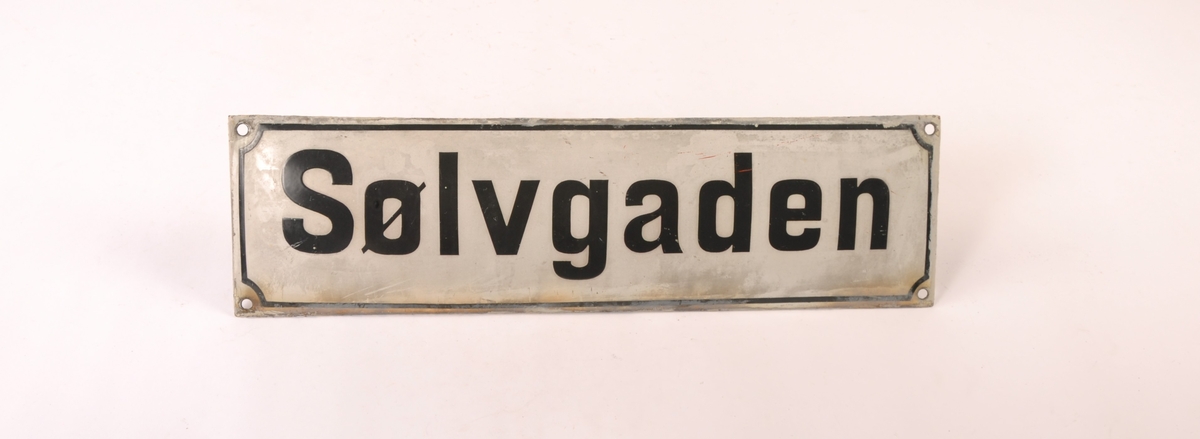 Rektangulært, konvekst, skilt av metall med festehull i hvert hjørne. Emaljert. Hvitt med sort tekst.