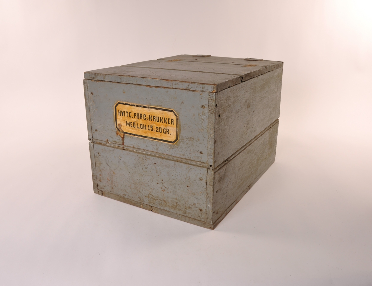 Rektangulær blågrå trekasse med flatt lokk med lær hengsler. Etikett, hvit med sort skrift. Ombygget lokk for større åpning.