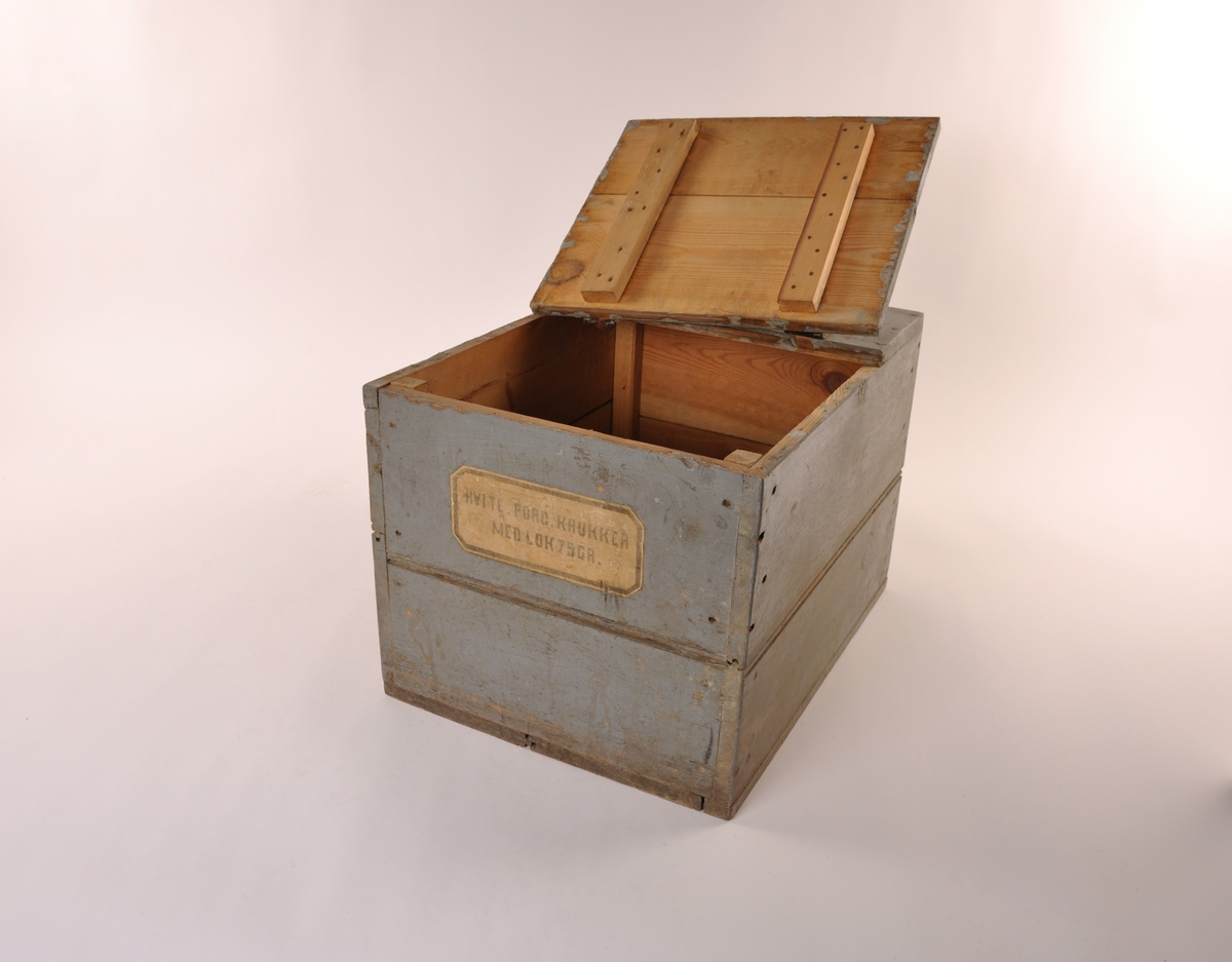 Rektangulær blågrå trekasse med flatt lokk med lær hengsler. Etikett, hvit med sort skrift.