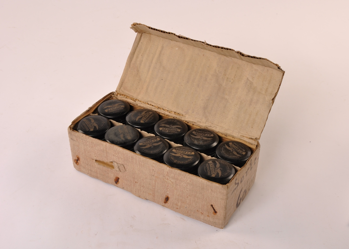 10 stk sylindriske krukker av sort porselen. Lokk av plast med apotekets logo i gull. I original embalasje av papp med årdring imitasjon