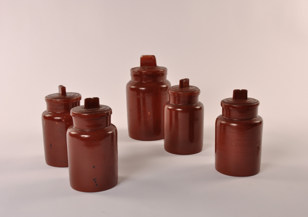 5 sylindriske glasskrukker med lokk. Malt med rød-brun farge på både innside og utside. 4 i lik størrelse + 1 som er større. Uten etikett. På innsiden av lokket: H. HEYE.