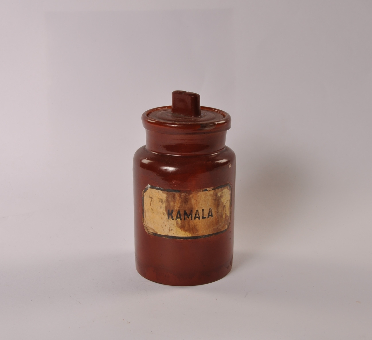 Sylindrisk glasskrukke med lokk. Malt med rød-brun farge på både innside og utside. Papiretikett med sort tekst limt på.