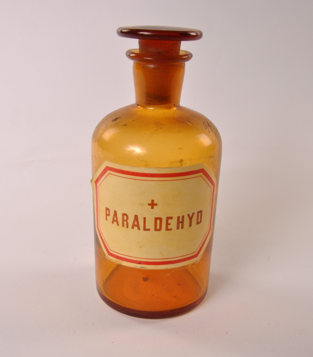 Sylindrisk glasskrukke i brunt glass med lokk. Påmalt etikett med sort skrift og ramme.