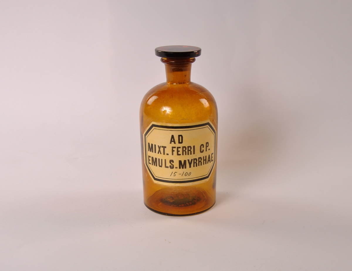 Sylindrisk glasskrukke i brunt glass med lokk. Påmalt etikett med sort skrift og ramme.