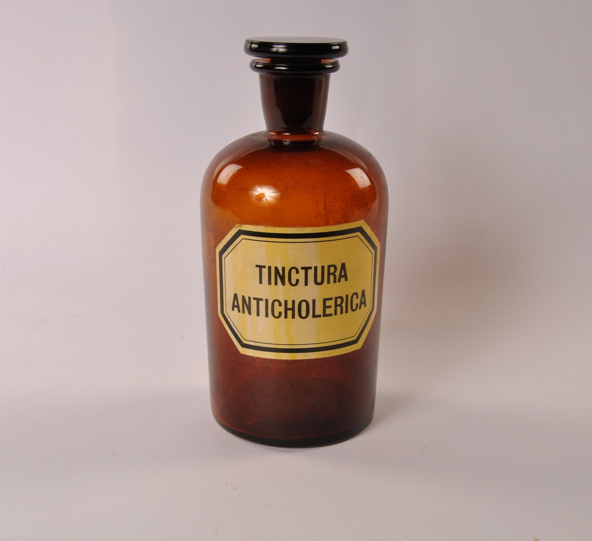 Sylindrisk glasskrukke i brunt glass med lokk. Påmalt etikett med sort skrift og ramme.