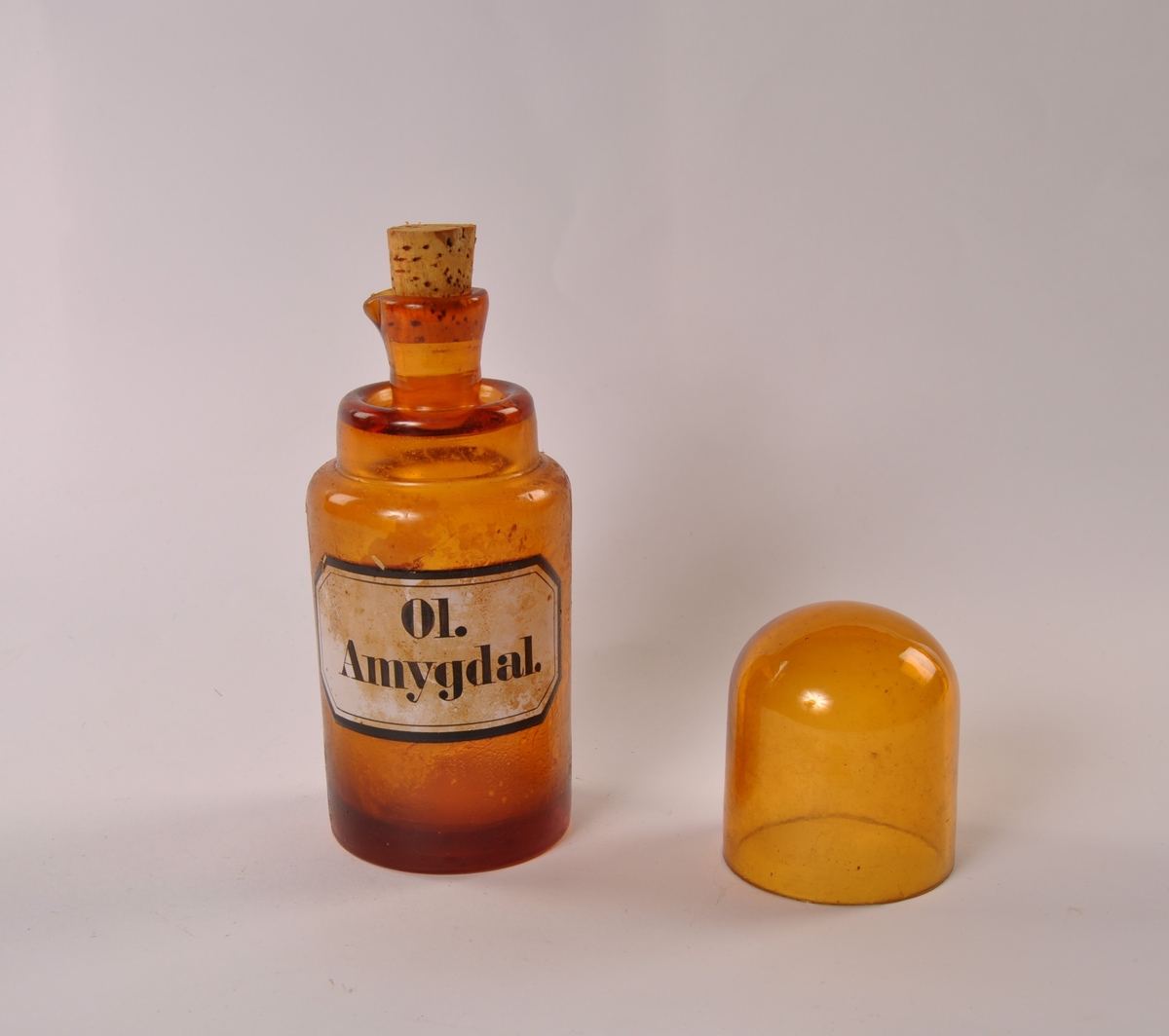 Flaske i brunt glass med rund hovedform, smalere hals med helletut. Kork. Rundtbunnet glasshette løst på toppen.