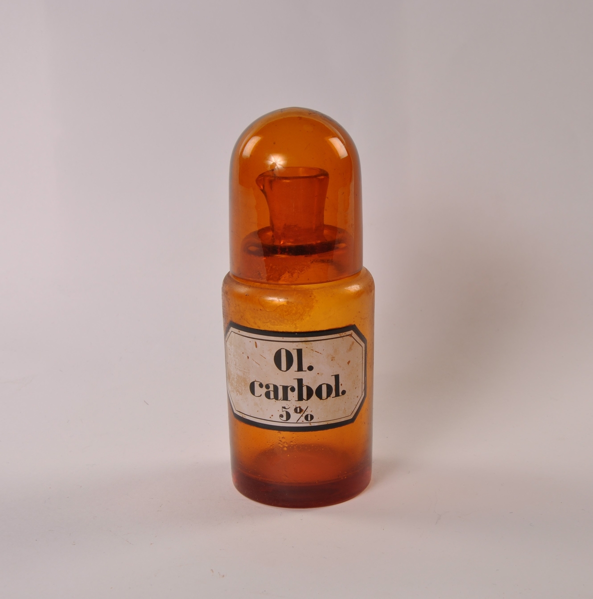 Flaske i brunt glass med rund hovedform, smalere hals med helletut. Rundtbunnet glasshette løst på toppen.