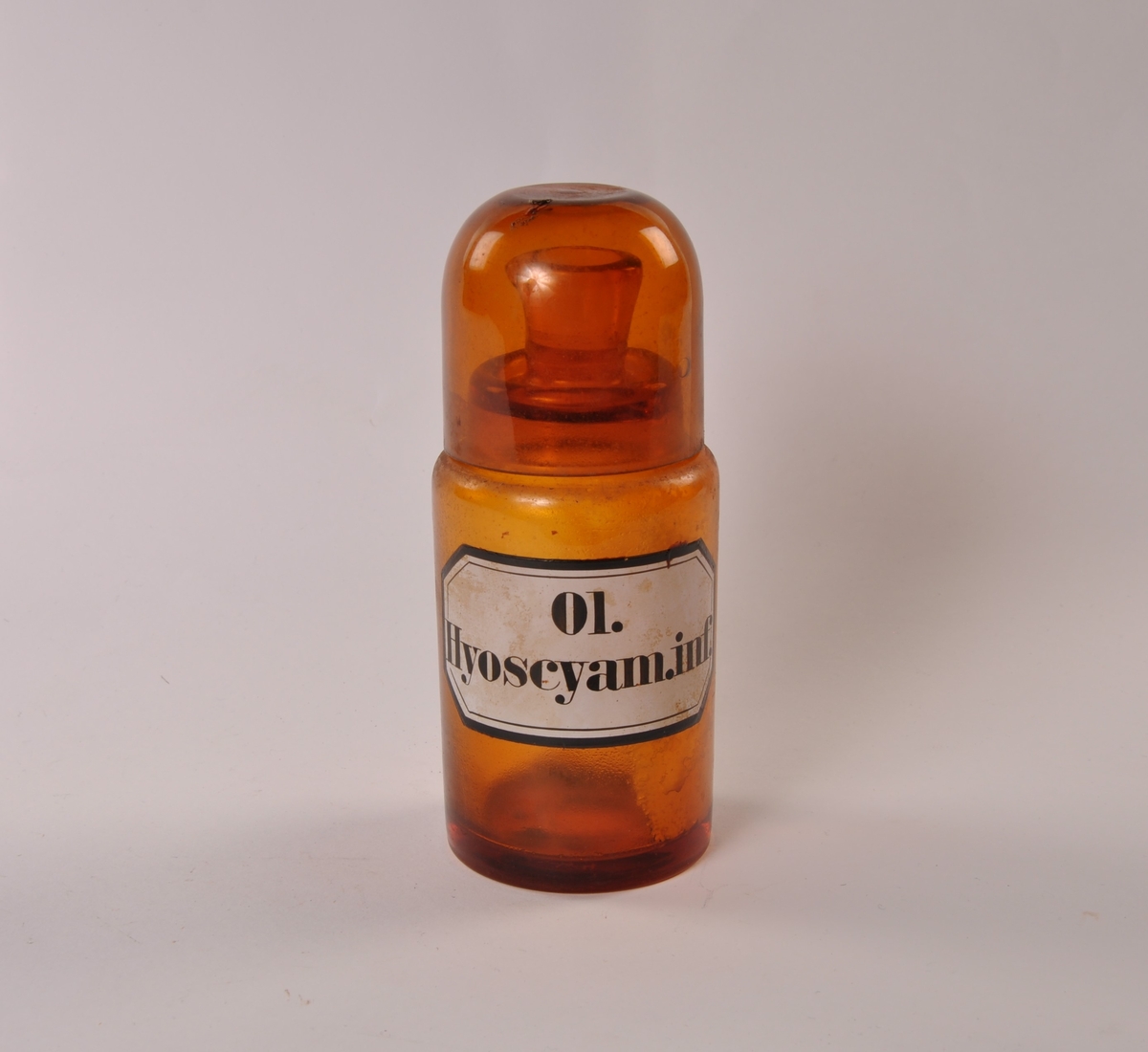 Flaske i brunt glass med rund hovedform, smalere hals med helletut. Rundtbunnet glasshette løst på toppen.