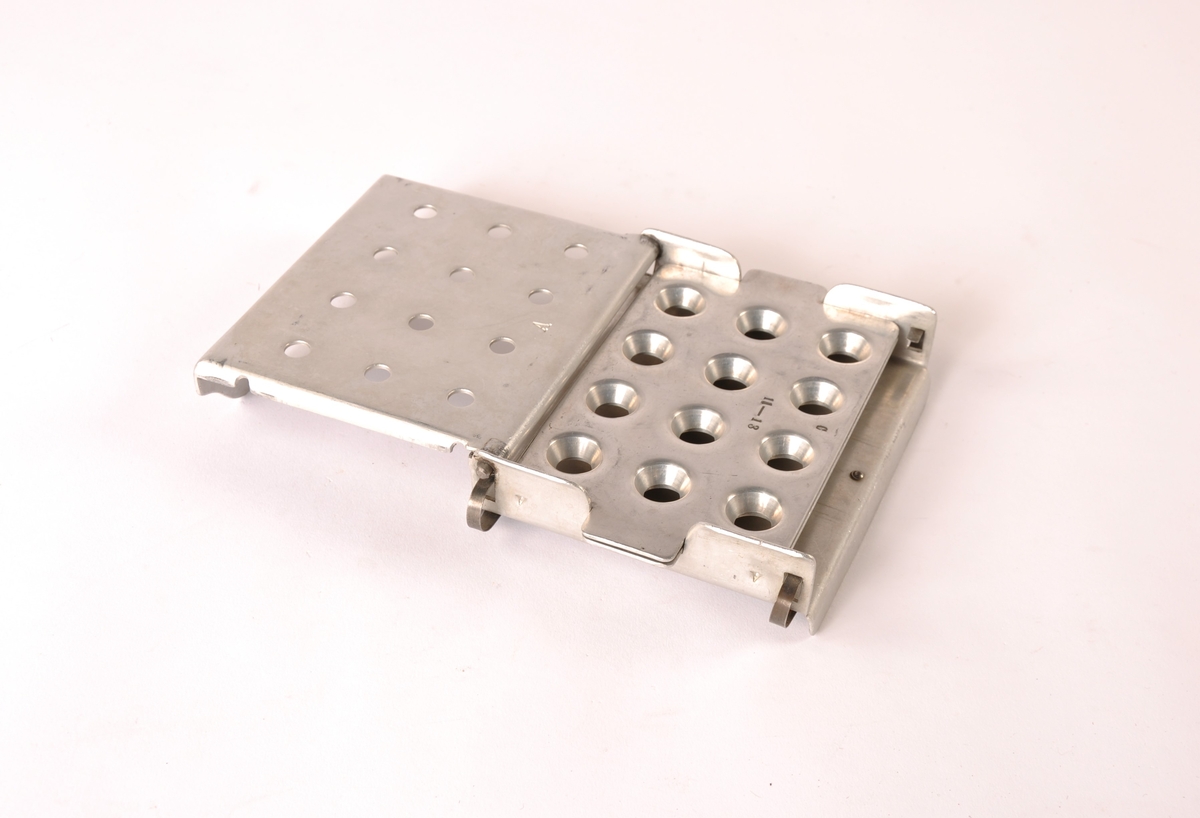 Forsegligsapparat i hvitt metall, trolig aluminium. To hengslede deler med kant og hull til pastillene.