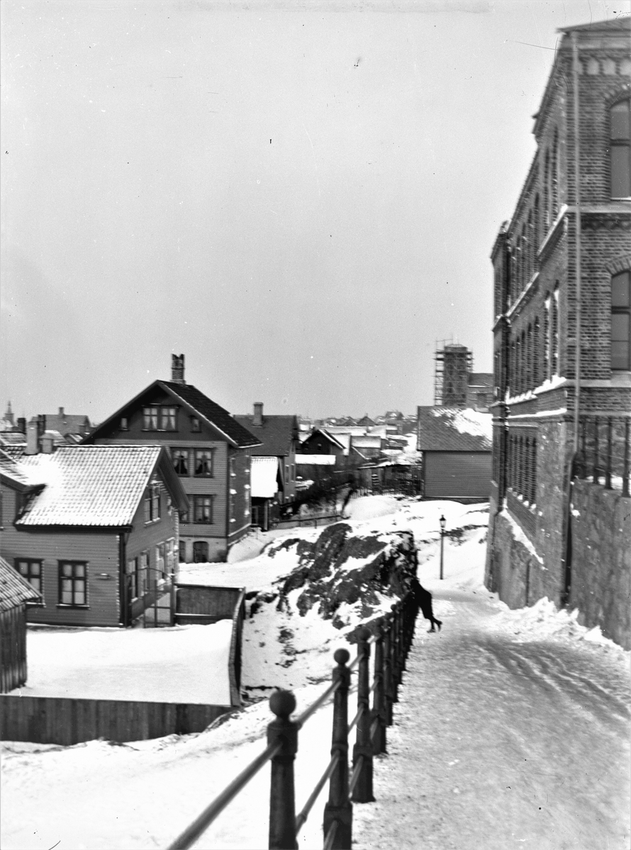 Parti av Haugesund. Bakken vest for Breidablikk skole. Rekkverk langs bakken. Skolen på høyre side. Vår Frelsers Kirke under konstruksjon. Byhus. Vinterbilde med snø.