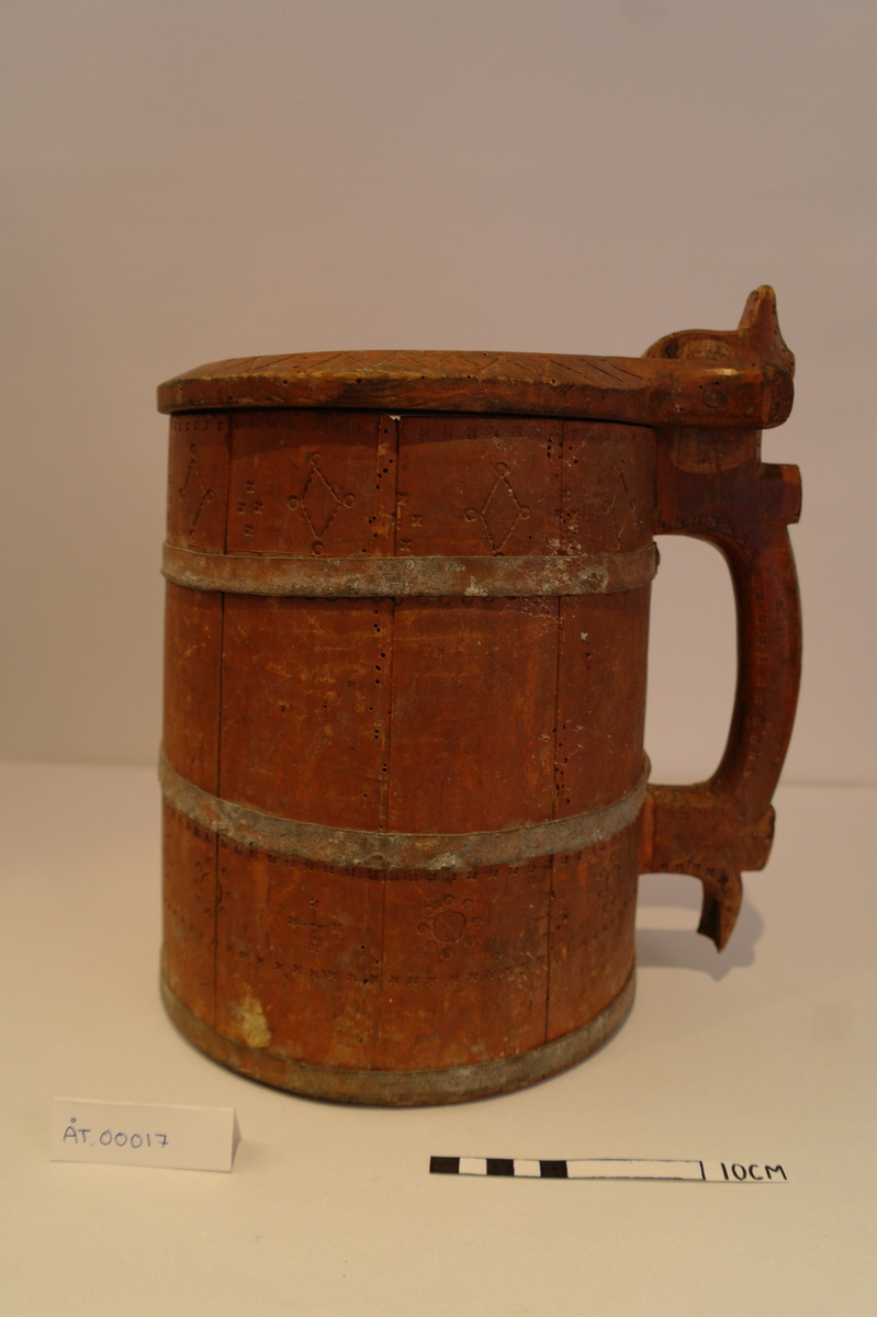 Lagga kar holdt saman av tre sinkband. Hanken er skrudd fast. Loket er hengsla til hanken med ein tretapp. Stavane, hanken og loket er dekorert med innrissa border,sirkler, spekulasjer og små blomer. Hanken er skåre ut i ein dekorativ fasong. Kanna er målt i ein raudbrun farge. I god stand. Nokre få hol etter mit.