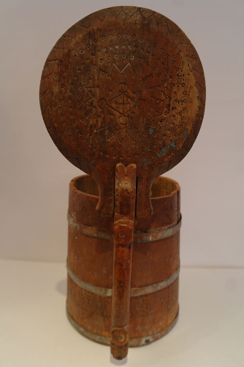 Lagga kar holdt saman av tre sinkband. Hanken er skrudd fast. Loket er hengsla til hanken med ein tretapp. Stavane, hanken og loket er dekorert med innrissa border,sirkler, spekulasjer og små blomer. Hanken er skåre ut i ein dekorativ fasong. Kanna er målt i ein raudbrun farge. I god stand. Nokre få hol etter mit.