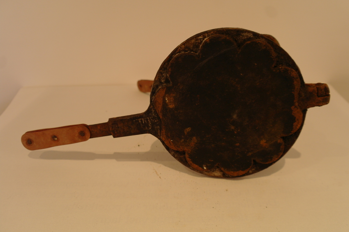 Vaffeljarn i jarn til bruk over vedovn. Produksjonsstempel:MUSTAD. På handtakene er det festa noko som liknar lær  med jarnnaglar. Innvendig er jarnet noko rusta.