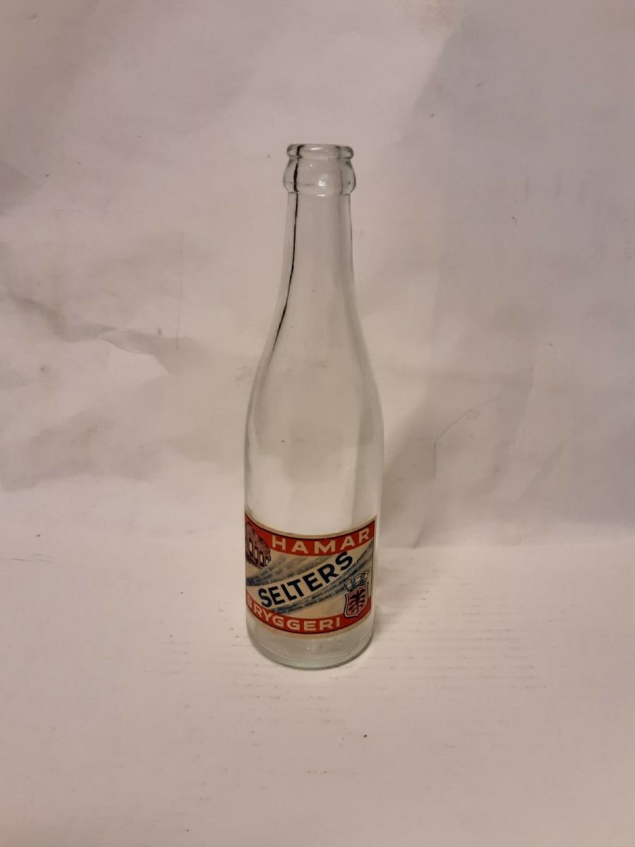 Seltersflaske, gjennomsiktig glass. Etikett av papir med tegning av domkirkeruinene og Hamars byvåpen. Tekst etikett: "HAMAR", "SELTERS", "BRYGGERI".