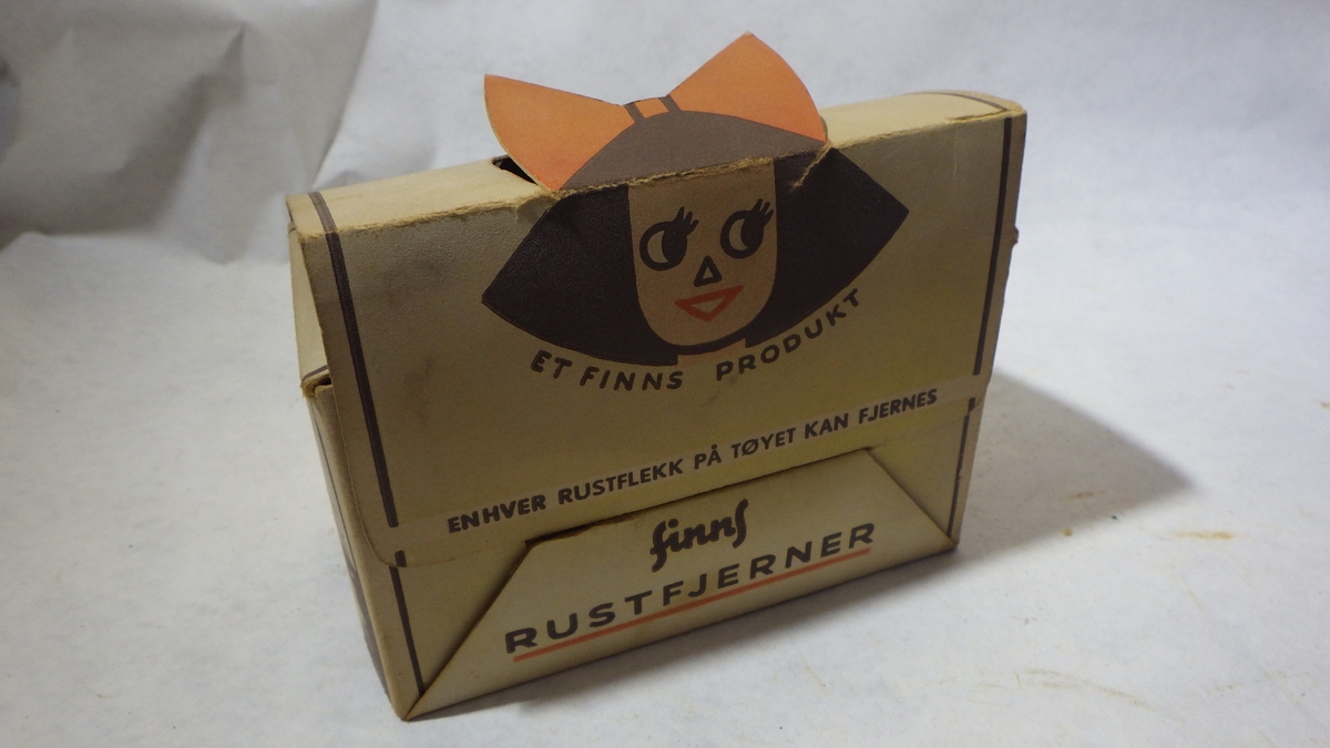 Pappeske gulhvit m/svart skrift. Tegning av ung kvinne med rød hårsløyfe, tekst: Et Finns produkt / Envher rustflekk på tøyet kan fjernes / Finns rustfjerner. I kartongen er det inndelt
for 24 tuber, 12 stk. er i esken. Tubene er grønne av plastikk. Rød merkelapp med varsel er
festet omkring med pakkestrikk, svart kork.
