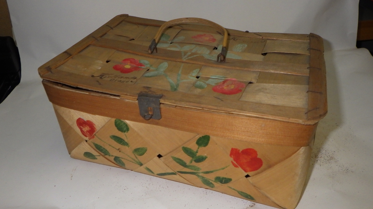 Flettet sponkurv med lokk. Blomsterkvister malt på sidene og på lokket. Liten trehank på lokket. Hekte av metall, som lukker og låser lokket. Tekst lokket: Kiellands Drage