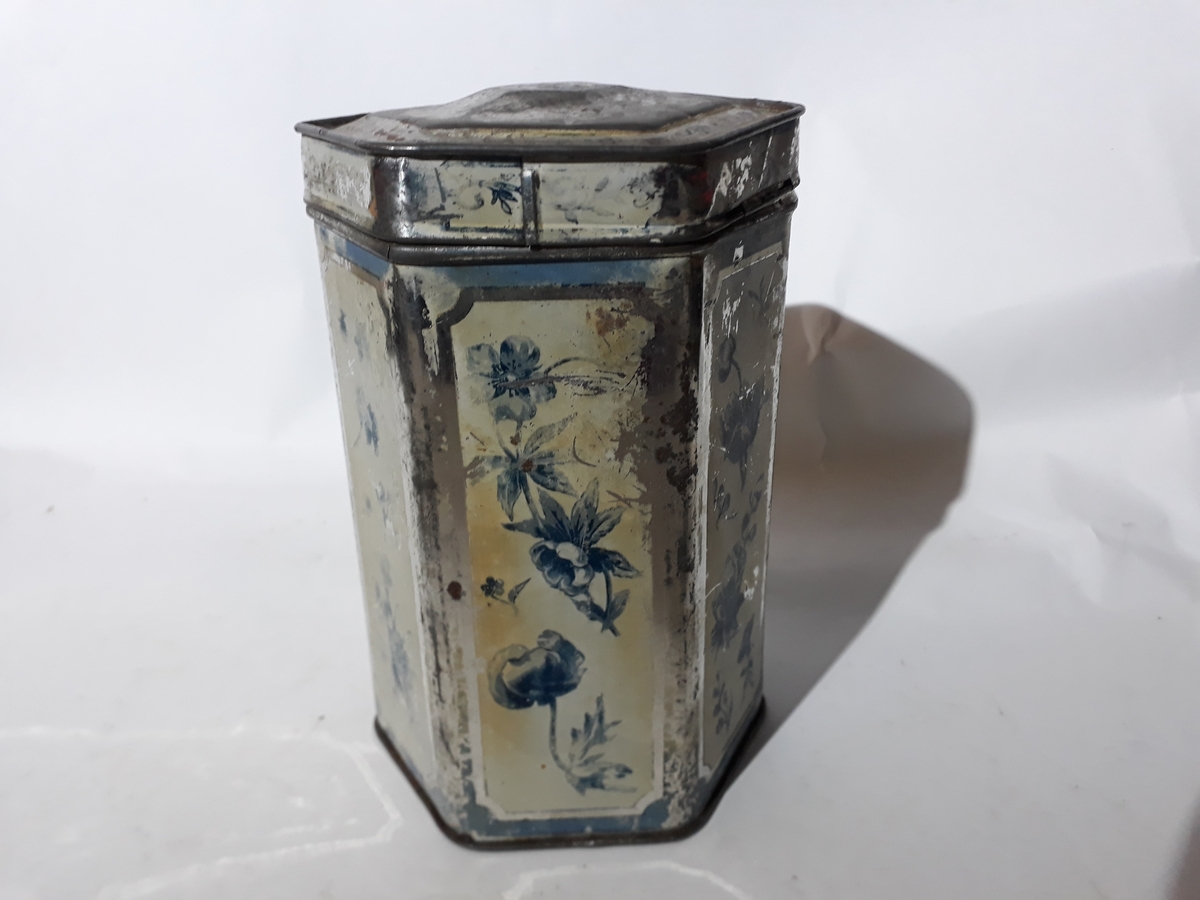 6-kantet blikkboks m/lokk. Hver kant har påmalt et felt med gul bunn og blå blomstermotiv.