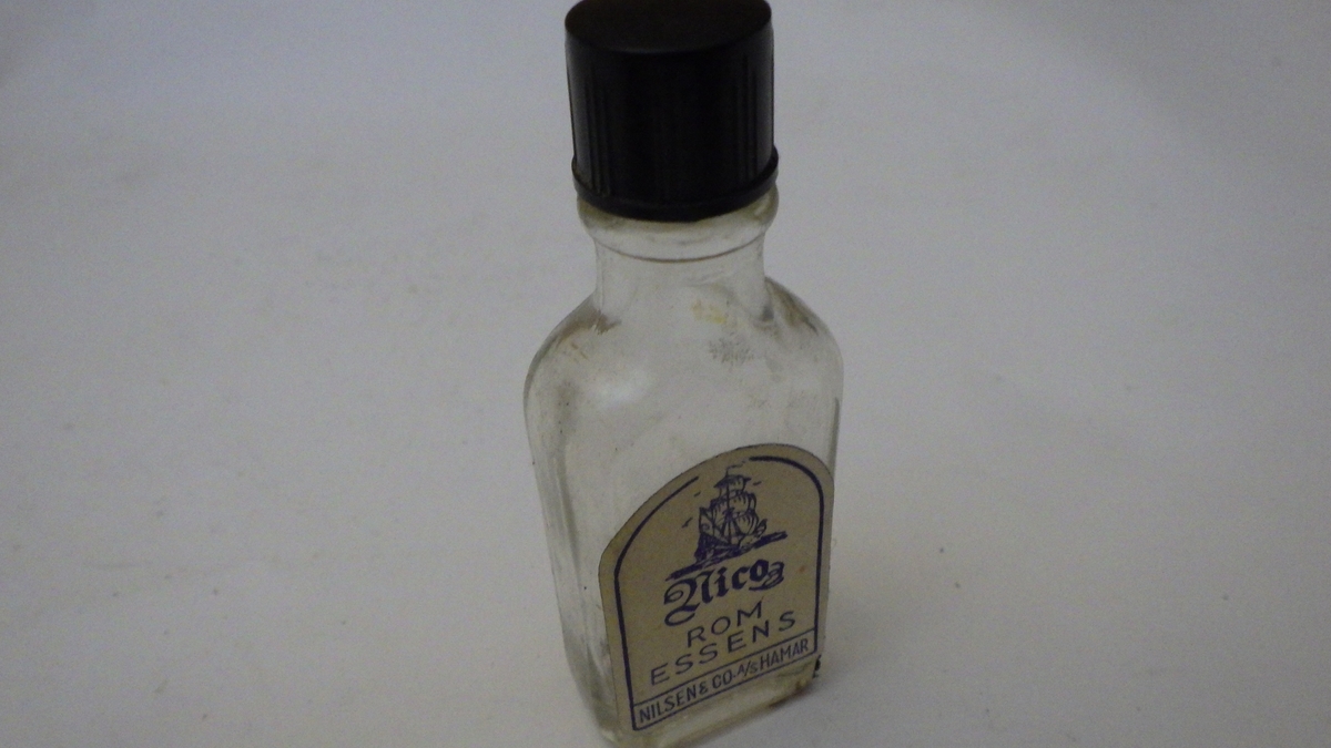 Flaske av klart glass, bakelitt skrukork. Etikett av papir, hvit bunn og blått trykk. Etter stilpreg fra 1950 årene(?).