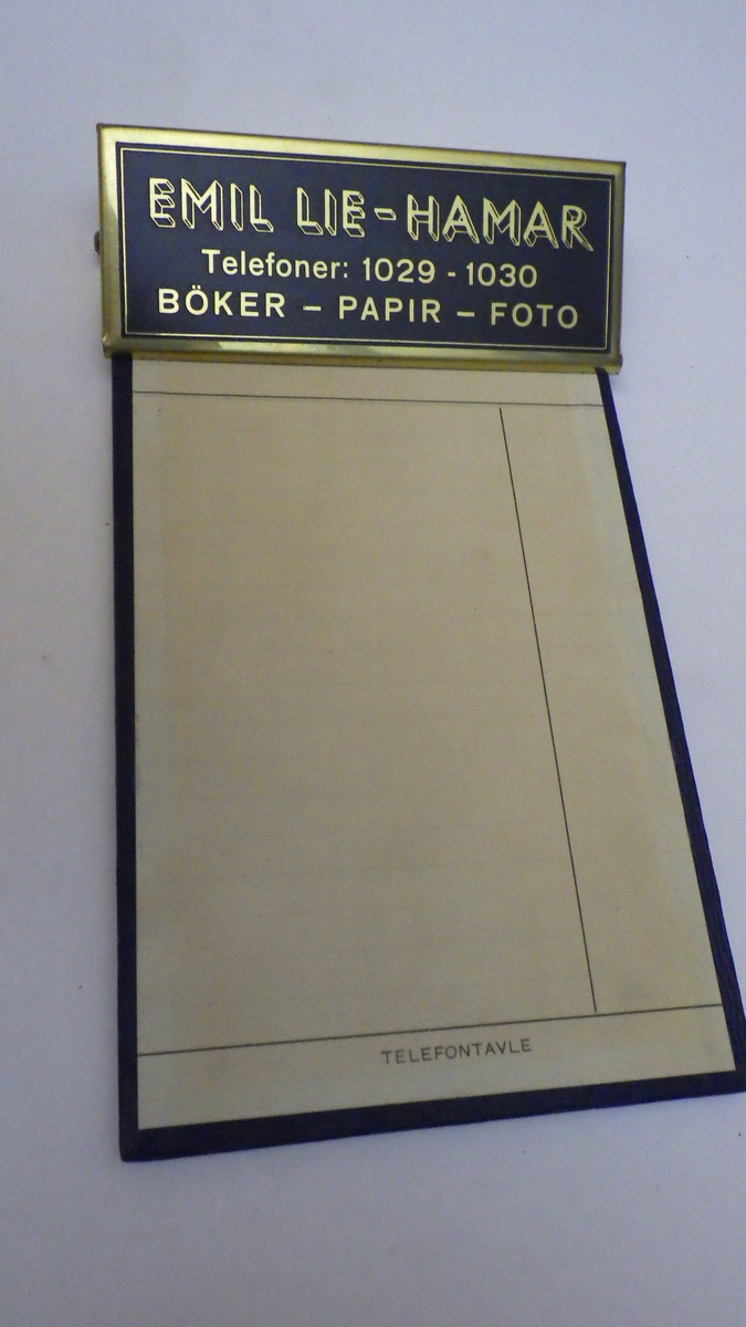 Plate av finer, trukket med papir. Klemme av metall, på sort bunn med tekst trykt i gull. Etter stilpreg fra 1930 -40 årene(?).