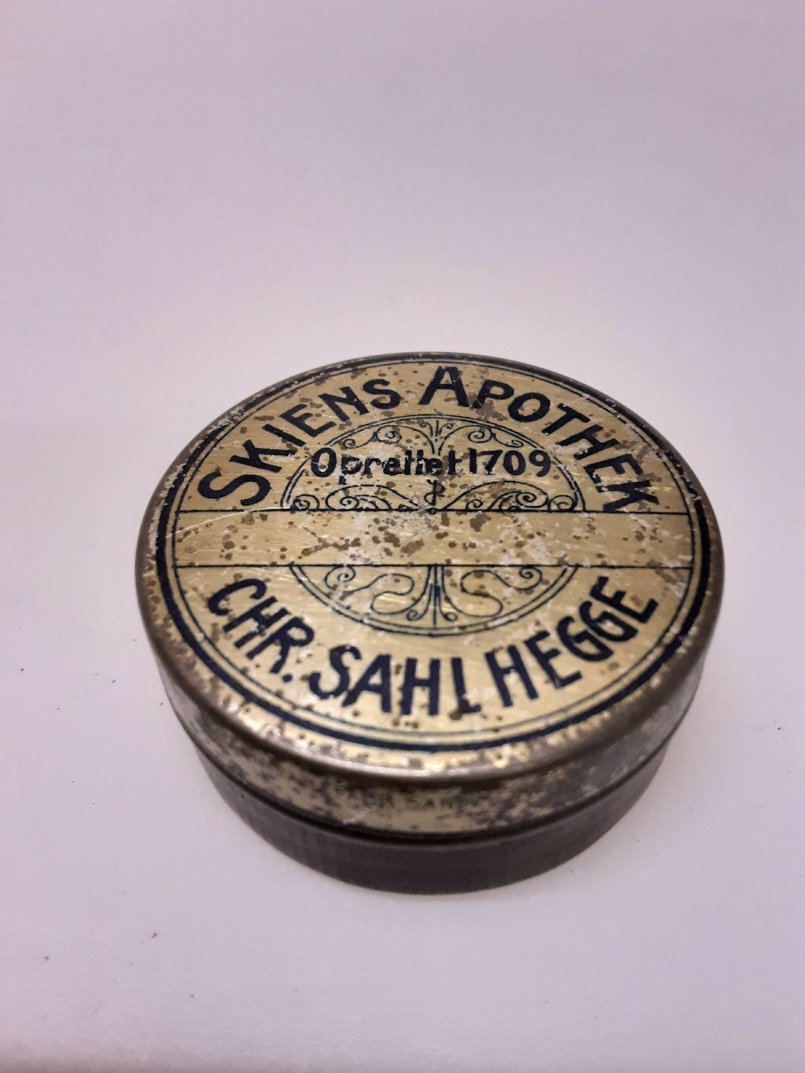 Blikk, eske med lokk, rund. Blå bokstaver, inskripsjon: «SKIENS APOTHEK OPRETTET 1709» / «CHR. SAHL HEGGE».
CHR.SAHI, HEGGE.