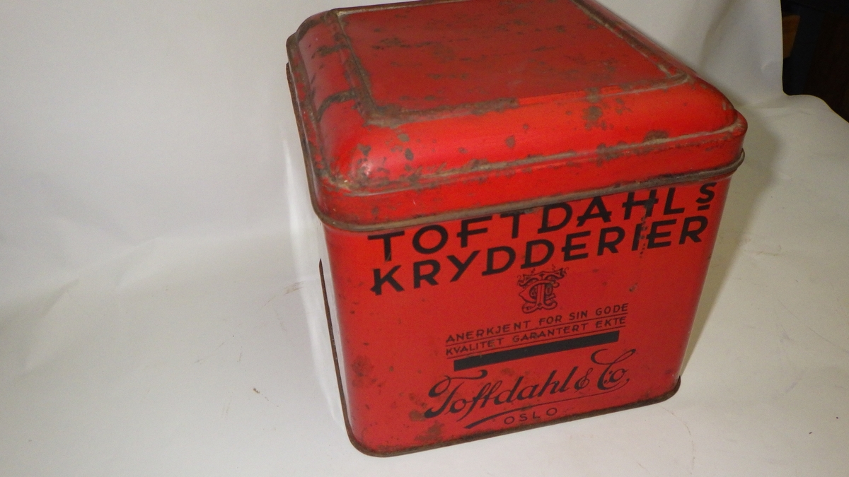 Blikk, kvadratisk, hengslet lokk. Rød, bokstaver i sort.