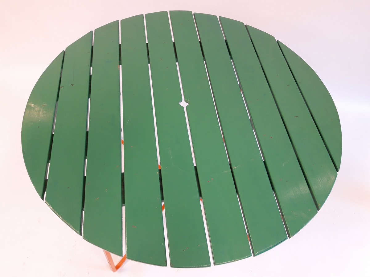 Ett rundt bord med 10 grønnmalte planker som bordplate. Understell av oransjemalt metall. Hull og feste for parasoll.  Stolene er laget av fjærstål som gir mulighet for svikt.