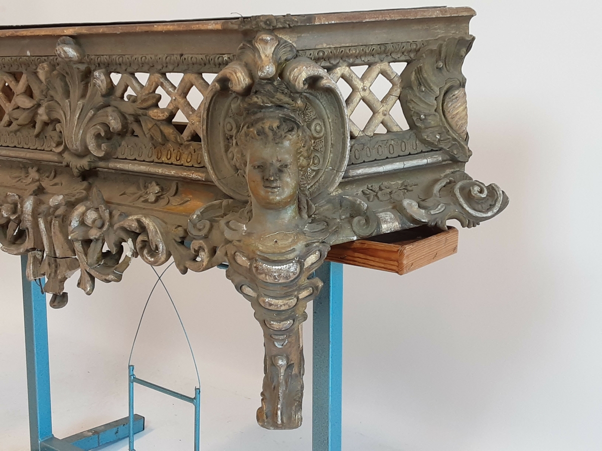 Lavt konsollbord med utskårne elementer; ett kvinnehode på hvert hjørne. Rik rokokkodekor som er forgyldt. Metallbakke nedfelt på toppen. Antagelig for blomsterdekorasjon.