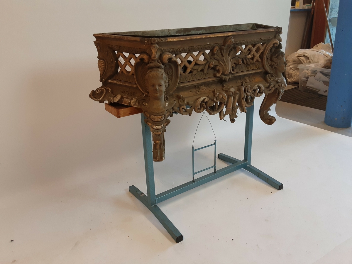 Lavt konsollbord med utskårne elementer; ett kvinnehode på hvert hjørne. Rik rokokkodekor som er forgyldt. Metallbakke nedfelt på toppen. Antagelig for blomsterdekorasjon.