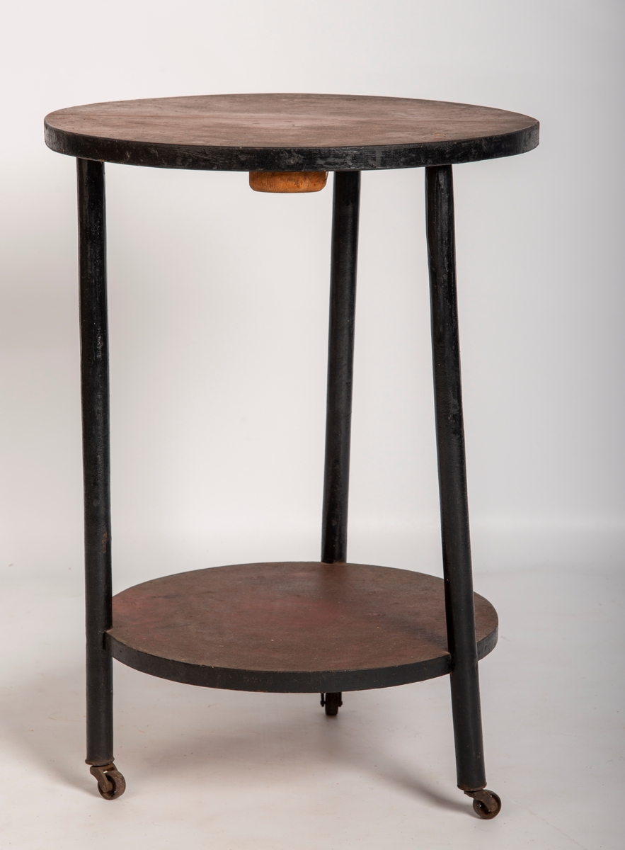Rundt bord med tre bein med hjul på. Hylleplate 17 cm. fra gulvet. Plater beiset i mørk rød farge. Bein og kanter på platene er malt/lakkert sorte.