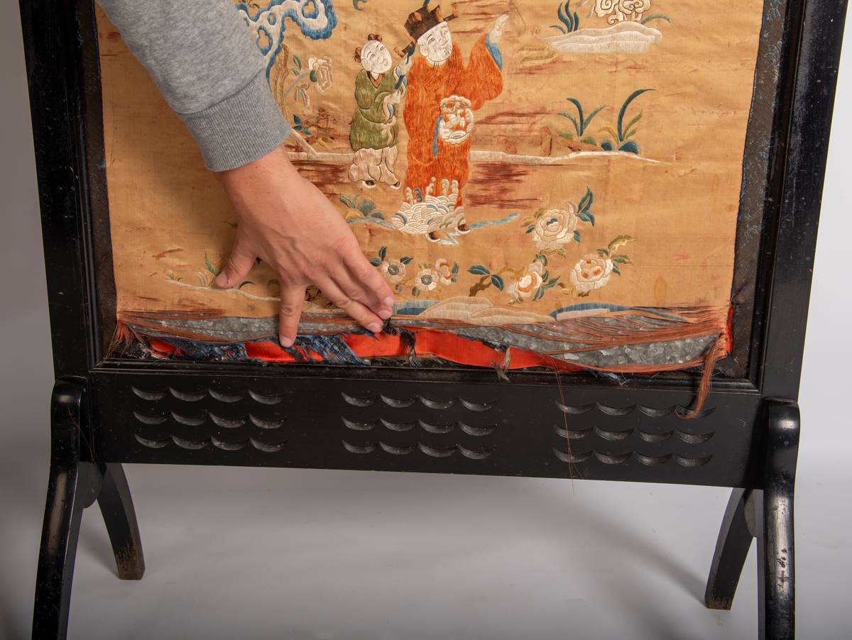 Svartlakkert ramme på 4 bein. Utskjæring av 6 blomstermotiv på toppstykket og 3 felt a 12 utskjærte bølger på nedre rammedel. Midtfeltet består av metallplate trukket med glanset bomull på baksiden og silkestoff med brodert asiatisk motiv på framsida.  2,5 cm bredt mønstret bånd limt på silkestoffet på alle 4 kanter., båndet består av en blanding av ull og silketråder.