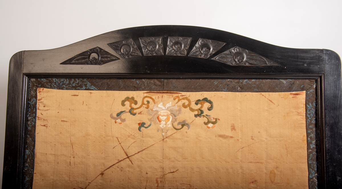 Svartlakkert ramme på 4 bein. Utskjæring av 6 blomstermotiv på toppstykket og 3 felt a 12 utskjærte bølger på nedre rammedel. Midtfeltet består av metallplate trukket med glanset bomull på baksiden og silkestoff med brodert asiatisk motiv på framsida.  2,5 cm bredt mønstret bånd limt på silkestoffet på alle 4 kanter., båndet består av en blanding av ull og silketråder.