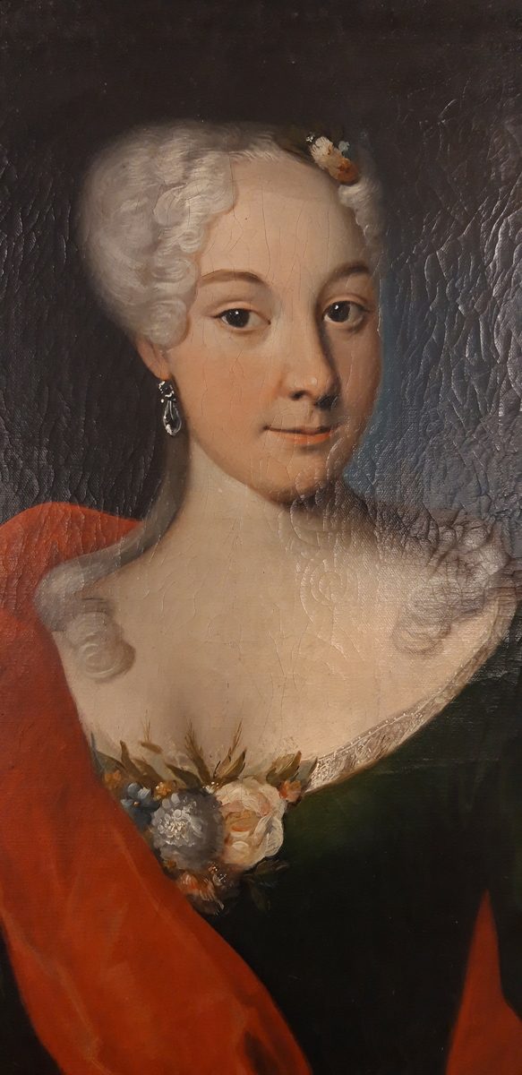 Portrett av ukjent kvinne, 1700-tallet, brystbilde, vendt svakt mot venstre. Trekvart profil og hodet er vendt høyre, med blikket mot betrakter. Mørk grønn utringet kjole med blondekant og blomsterdekorasjon. Øredobb i hennes høyre øre. Lyst oppsatt hår, lange lokker over skuldrene og hårpynt av blomster. Rødt sjal er drapert over kvinnens høyre skulder og venstre arm. Blålig felt i bakgrunnen på høyre side og det er malt en mørk oval ramme rundt motivet.