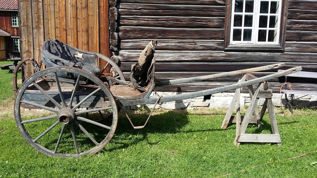 Vogn med to hjul, flatbunnet med c-fjærsopphengt sete (ett tverrstilt), skinntrukket fløy på jernramme som hovedveske (?), skvettlær.
Greaseaksel.
Jernbeslåtte hjul.
Antall trefelll: 6
Antall eiker: 12