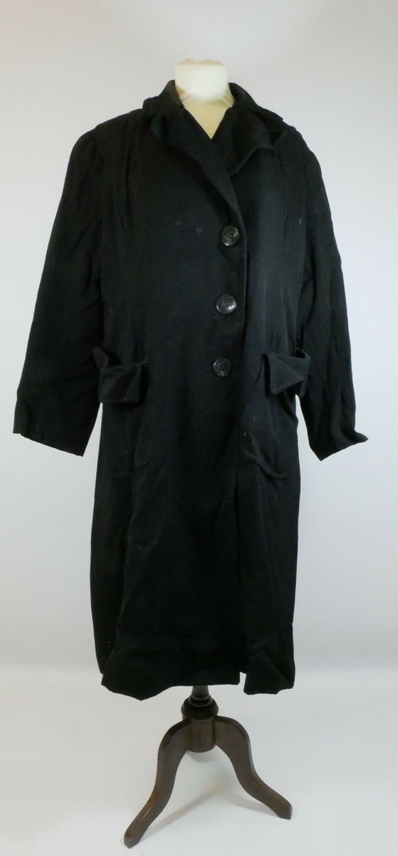 Svart kåpe med krager og ermer. Kåpen har enkeltspenn med tre svarte runde knapper foran. Kåpa er foret og har to store lommer. 