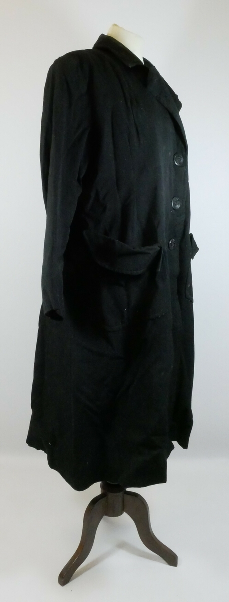 Svart kåpe med krager og ermer. Kåpen har enkeltspenn med tre svarte runde knapper foran. Kåpa er foret og har to store lommer. 
