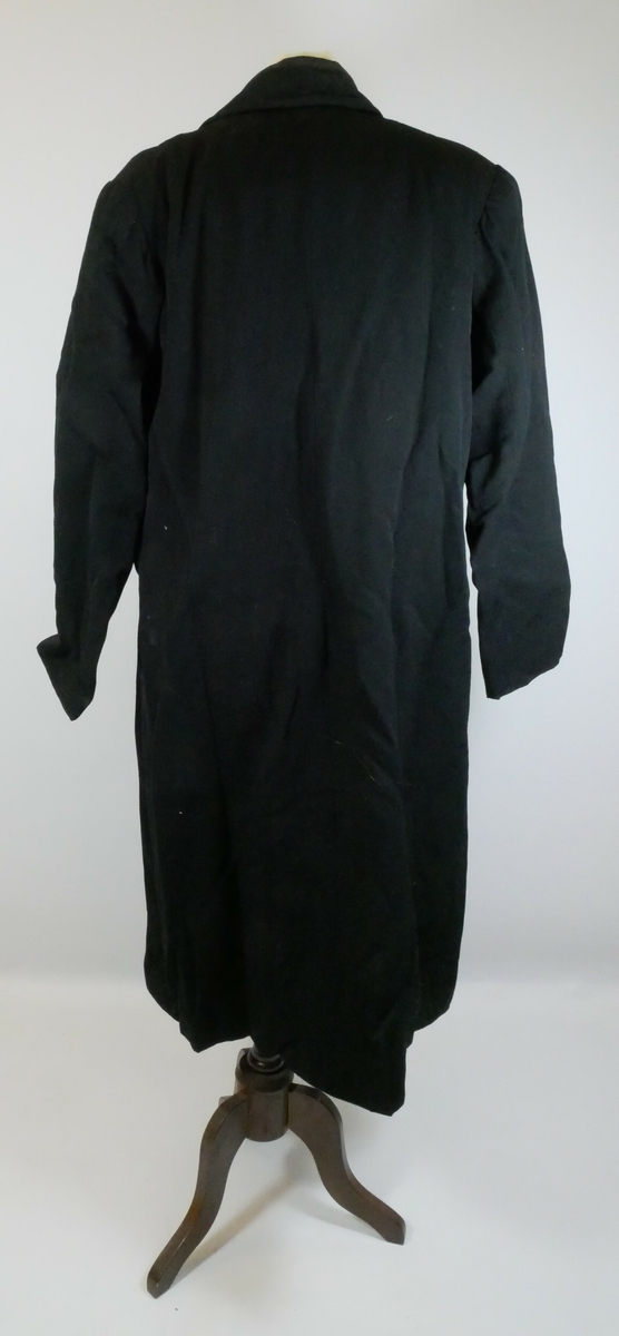 Svart kåpe med krager og ermer. Kåpen har enkeltspenn med tre svarte runde knapper foran. Kåpa er foret og har to store lommer. 