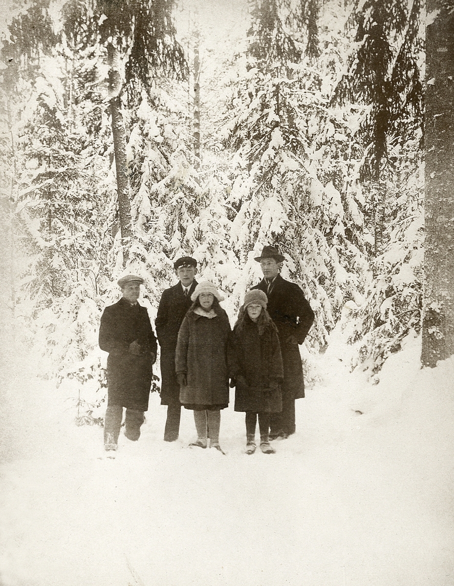 Två män, en halvvuxen pojke i skolmössa och två flickor poserar för fotografen i en vintrig skog. 
Text under fotot: "Skogen i vinterskrud, 1923".