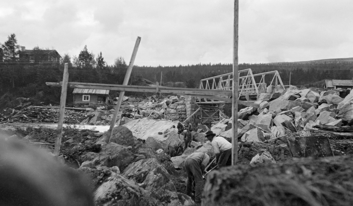 Atnsjødammen under bygging vinteren 1916-17.  Fotografiet er tatt fra elvas østre bredd, der flere karer arbeider blant store steinblokker som antakelig skal brukes i damkonstruksjonen.  I forgrunnen er det montert en slags «portal» av bjelker, muligens som en markering av det som etter hvert skal bil damarmens tverrsnitt.  Til høyre ses ei vegbru av støpejern.  Brua er bygd i ett spenn mellom landkar av hoggen naturstein.  Et intakt tresillas under det vestre brukaret tyder på at også denne konstruksjonen var forholdsvis ny da fotografiet ble tatt.  På vestsida av elveløpet ligger ei lita laftet bu på elvebakken, muligens ei damkoie.  På bakkekammen lengre bak, til venstre i bildet, ses en noe større stuebygning. Det statlige skogvesenet bidro med 5 000 kroner til dette dambyggingsprosjektet under forutsetning av at etatens tømmer fra området for ettertida ikke skulle belastes med damavgift. Denne fløtingsdammen skulle gi en reguleringshøyde på inntil 2 meter.