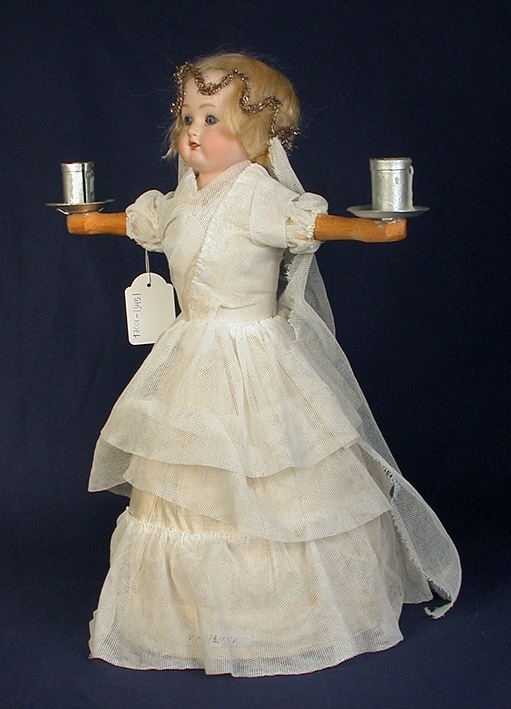 Lucian är klädd i en vitfärgad tyllklänning med tre volanger. Har träarmar som håller i var sin ljushållare. På huvudet har hon en slöja med en glittergirland runt håret.