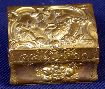 Av koppar, rektangulär form, underdel med lock som öppnas och stängs med hjälp av ett gångjärn i metall. Locket något välvt vilket gör att asken liknar en kista. Hela askens utsida dekorerad med mönster och figurer i relief; på lockets ovansida en man och en kvinna med utseenden som påminner om grekiska antiken, med varsitt instrument, harpa respektive något som liknar ett horn (?), i en ram av bladverk och kolonner.