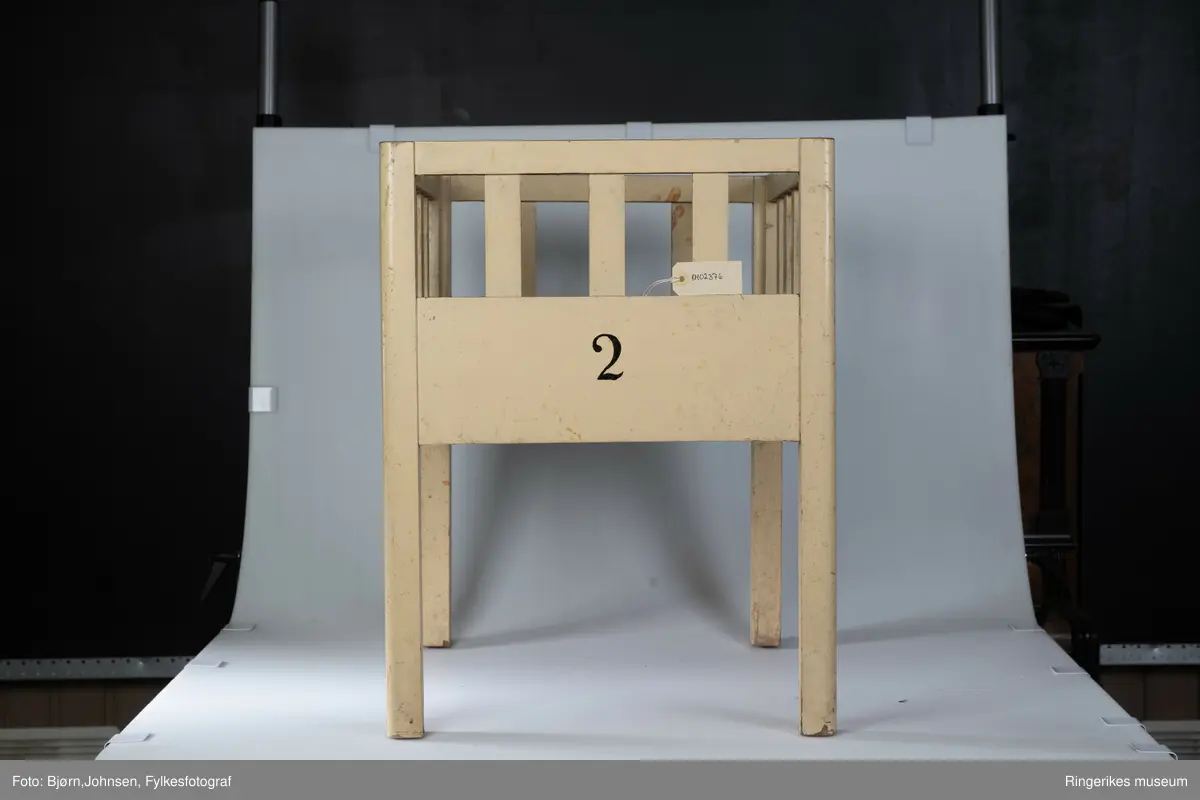 Gulmalt babyseng i tre. Tallet 2 er malt med svart på den ene kortsiden. Nederste del av senga er tett, øverste del har spiler, tre i hver kortside og fem i hver langside.