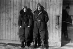 Grupporträtt av två militärflygare i flygoverall ståendes framför en hangarport, vintertid. I bakgrunden syns en annan man inne i hangaren. 1930-tal.