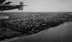 Flygbild av strandlinje vid stad. Till vänster syns två flygplan S 5 flygandes i roteformation. 1930-tal.