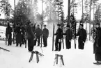 Flygsoldater står i en skog med skidor i hand, vintertid. Omkring tolv män tittar på en soldat. 1930-tal.