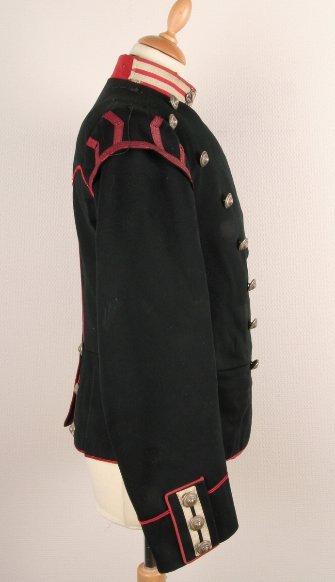 Offiserjakke fra den Norske Hæren. Svart jakke med røde og hvite dekorstriper under epaulettene. Stripene forestiller "svalereder" og lyrer på halskragen. Dette tyder på at jakka er uniform for en militær musiker. 14 sølvfargende knapper foran (to rader med 7 knapper) og 3 knapper på hvert erme. Knappene har diam: 23 mm. Jakka er norskprodusert.
Epaulettene på bilde 1 tilhører uniformen - er registrert som TM-12259